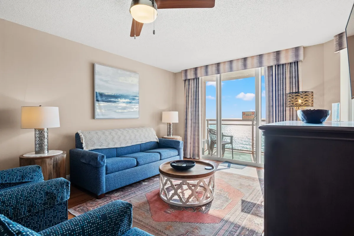 The image showcases a bright, airy living room with a plush blue sofa and stylish wooden coffee table. Large windows frame sweeping ocean views, allowing sunlight to fill the space. Artwork on the wall and an inviting balcony hint at relaxing mornings watching the sunrise and cozy evenings spent inside.