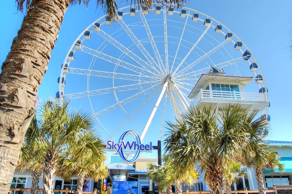 A vibrant exterior shot of the SkyWheel, framed by palm trees and welcoming entrance signage. The scene is lively yet romantic, suggesting fun shared adventures and memorable moments, with hints of nearby dining and attractions.