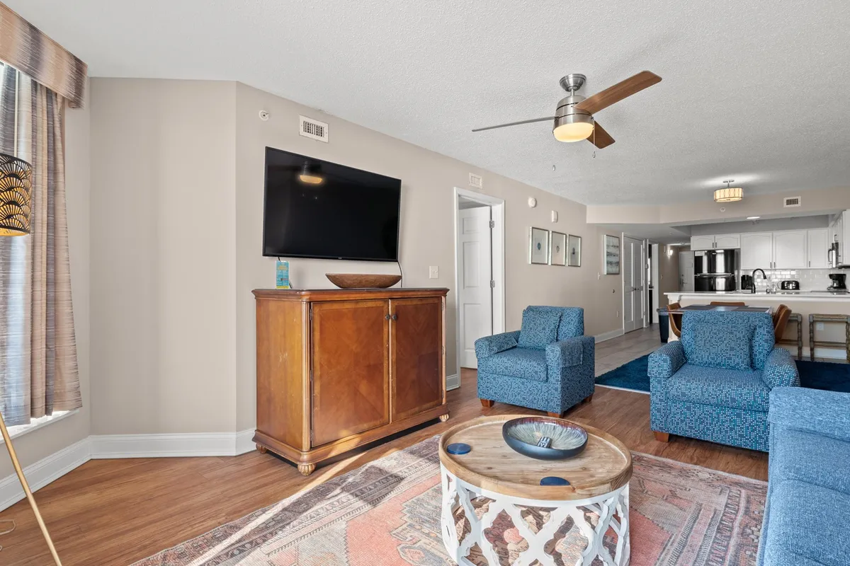 The photo reveals a cozy living room scene with a wooden cabinet, television, and a comfortable seating area. Large windows with curtains let in plenty of natural light, making the room feel spacious and inviting for relaxing or socializing.