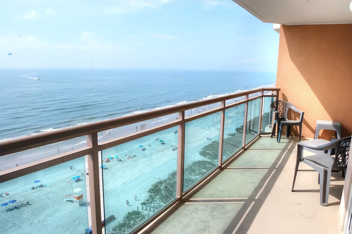 A balcony overlooking the beach with ocean views, beach umbrellas, and people enjoying the sun and sand. The scene captures the connection between the resort and the beach, emphasizing direct access and the lively coastal atmosphere.