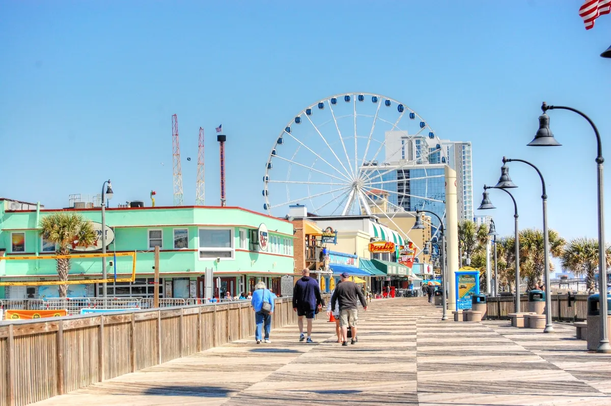 The image showcases the iconic Myrtle Beach SkyWheel, surrounded by the bustling boardwalk with shops and people walking along. The scene is lively and joyful, capturing the vibrant energy of one of the area's top attractions, especially during sunset.