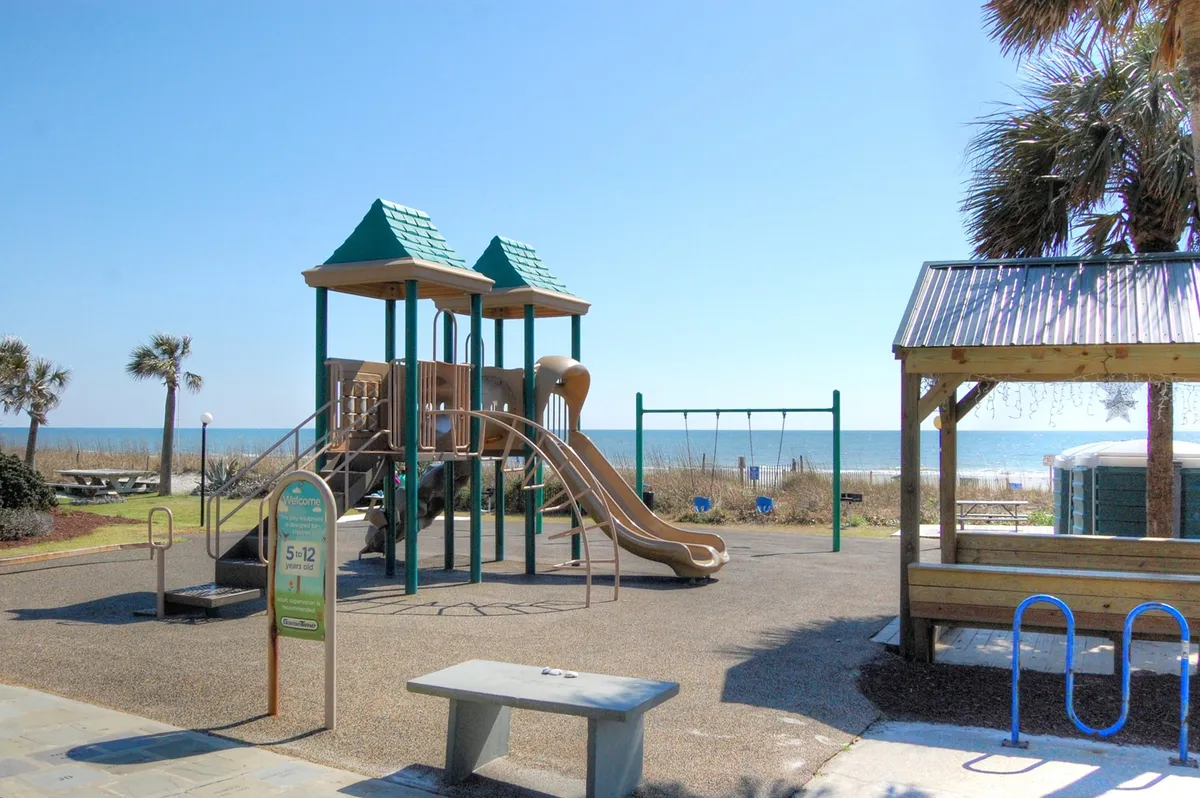 A colorful playground and picnic area set against a backdrop of beach views, with children playing and families relaxing. The scene is lively and joyful, highlighting kid-friendly amenities and outdoor fun.