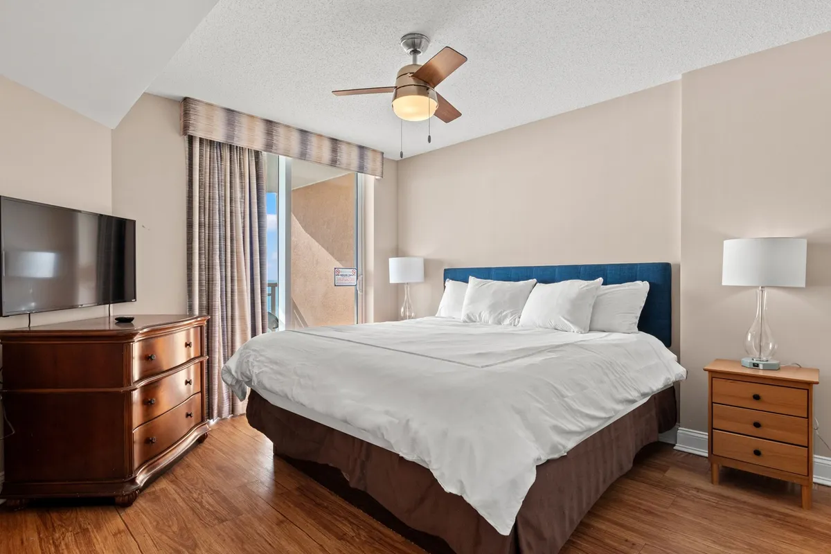 A serene bedroom featuring a neatly made bed with crisp white linens, matching nightstands with lamps, a television mounted on a wooden dresser, and soft curtains letting in gentle natural light. The room exudes comfort with cozy decor and ambient lighting.