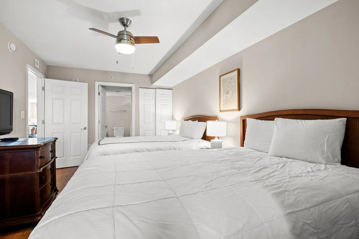 A spacious bedroom with two queen-sized beds, each adorned with inviting linens. The room includes a wooden dresser with a TV on top, matching table lamps, and framed artwork, creating an atmosphere of comfort and warmth—ideal for families or groups.