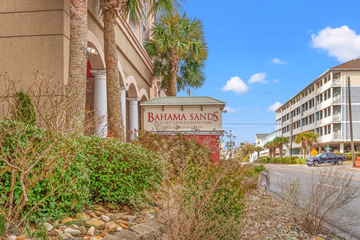 A welcoming exterior view of the Bahama Sands building, surrounded by vibrant palm trees and lush landscaping. The property's sign is clearly visible, and the atmosphere is warm and inviting, hinting at proximity to the beach and relaxing outdoor spaces.
