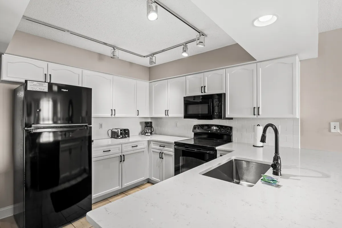 This image showcases a spacious, light-filled kitchen featuring white cabinetry, black appliances, and a large countertop. The layout suggests plenty of workspace for cooking and preparing meals, making it ideal for both casual breakfasts and more elaborate dinners. The surrounding area is open, hinting at easy access to adjacent living and dining spaces.
