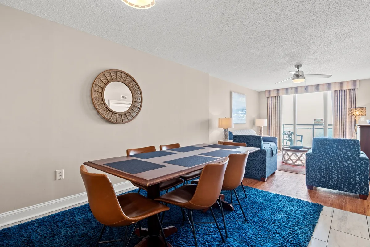 This image captures the inviting dining area with a dining table set atop a blue rug, a large mirror reflecting the light, and an open view into the adjoining living area. The soft, ambient lighting and visible balcony access highlight the seamless flow between spaces.