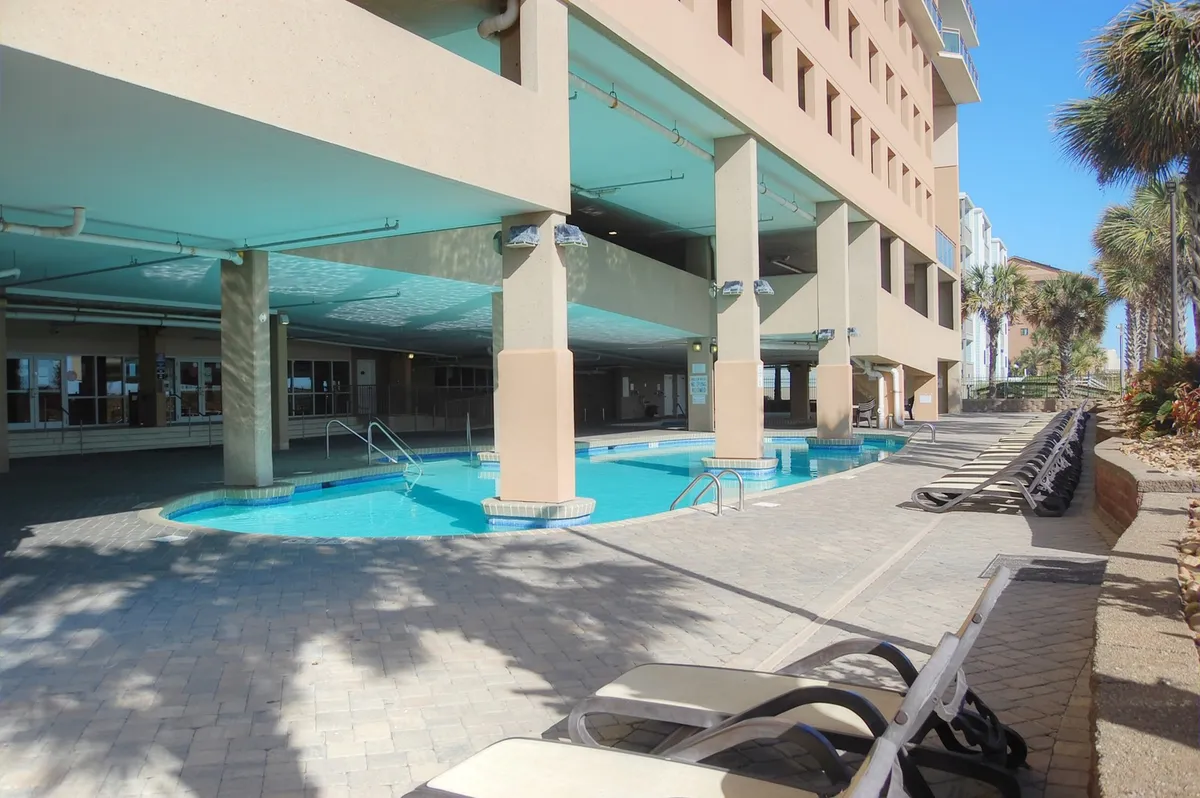 An inviting outdoor swimming pool surrounded by lounge chairs and palm trees, with the resort building visible in the background. The pool area is designed for relaxation and socializing and offers a tranquil ambiance ideal for soaking up the sun.