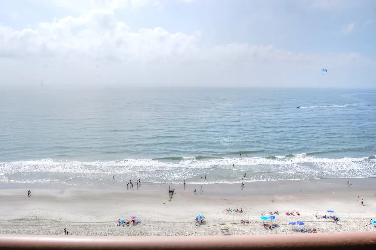 This vibrant image shows the shoreline bustling with activity: people are walking along the sand, beach umbrellas are set up, and parasailers soar over the gentle ocean waves. The scene radiates a joyful, carefree energy ideal for families enjoying the beach together.